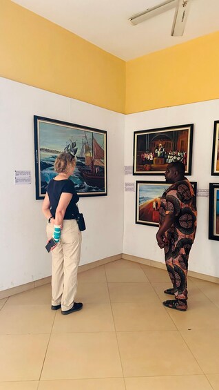 Two people examining framed artworks in a gallery with yellow walls and a tiled floor. 2