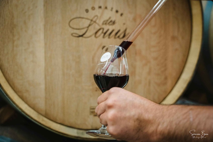 A hand holding a wine glass with a corkscrew, in front of a wooden barrel with 'Domaine des Louis' written on it. 3