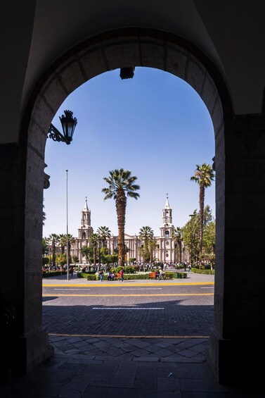 An archway framing a plaza with palm trees and a historic building with two towers. 1