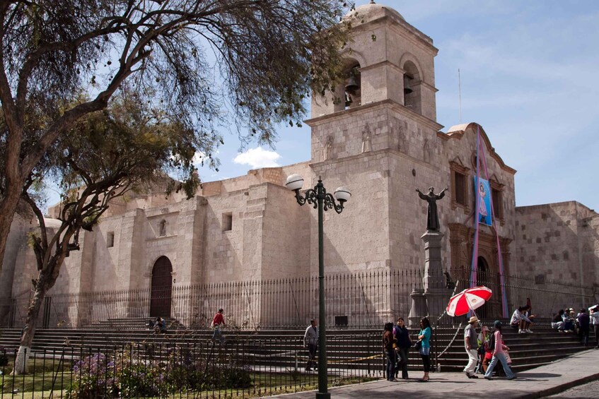 A historic stone building with a bell tower, surrounded by a fence and people walking. 2