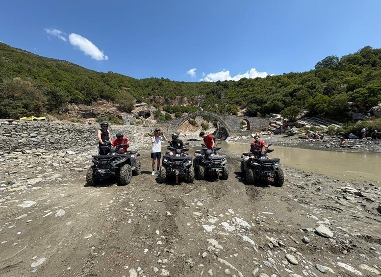 A group of people on ATVs near a rocky riverbank with a bridge in the background. 4