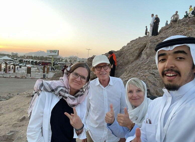 A group of people at a desert location with a rocky hill and a parking area in the background. 3