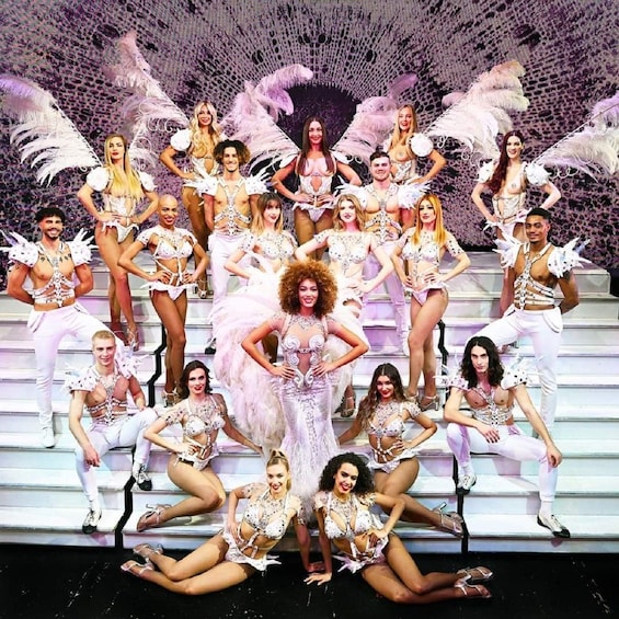 A group of performers on stage with feathered costumes and a backdrop of a patterned design. 1