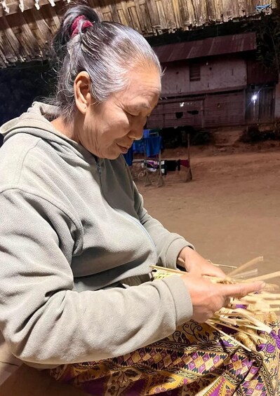 An elderly person seated outdoors, working with wooden sticks, possibly crafting or preparing them. 2