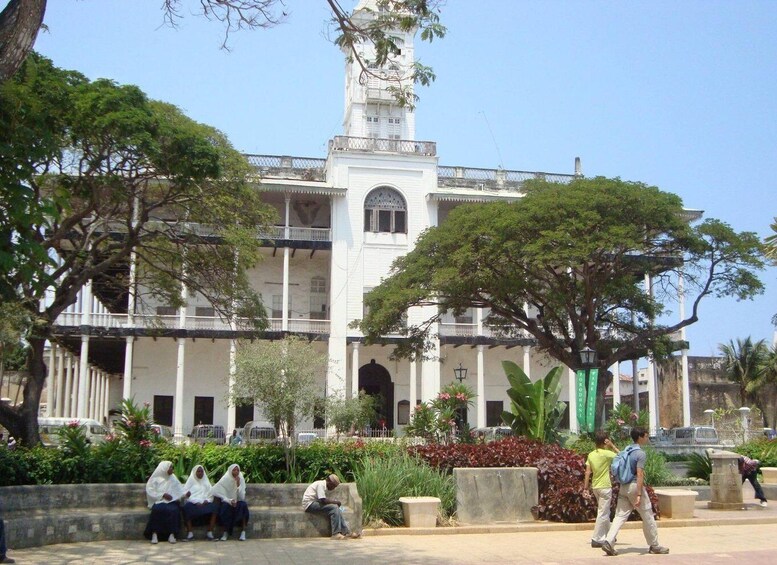 A large white building with a clock tower, surrounded by trees and people sitting on benches. 1