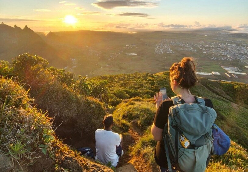 A person with a backpack and a water bottle sitting on a grassy hill overlooking a valley with a sunset in the background. 5