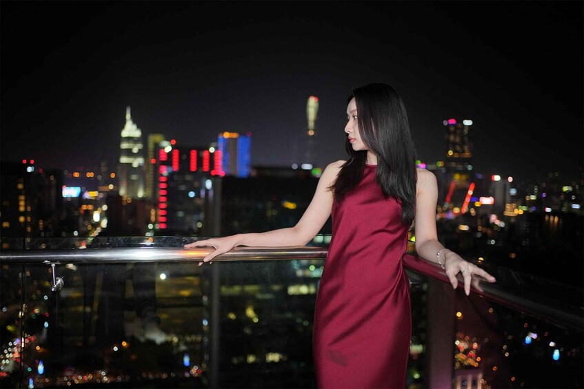 A person in a red dress standing on a balcony with a cityscape at night in the background. 2