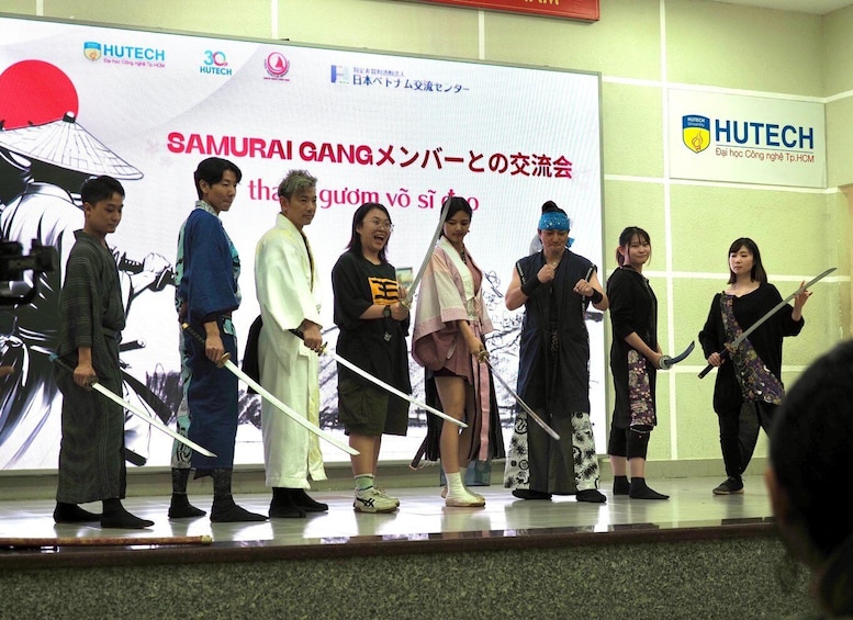 A group of people on a stage with swords, standing in front of a banner with Japanese text and logos. 4