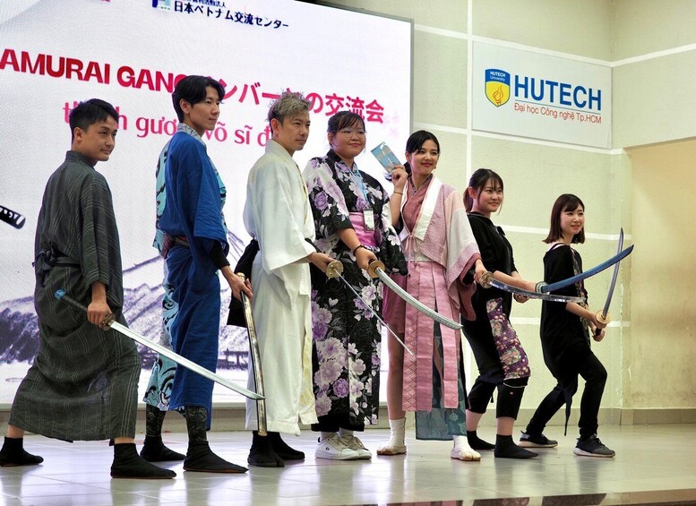 A group of people in traditional Japanese attire holding swords, standing in front of a banner with Japanese text and a HUTECH University logo. 5