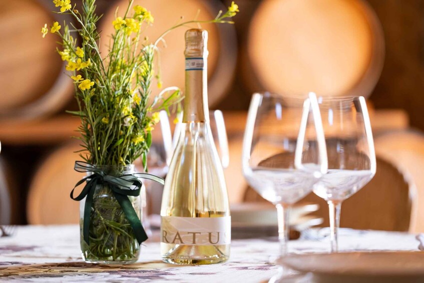 A bottle of white wine with a label, a glass vase with yellow flowers, and two empty wine glasses on a table. 2