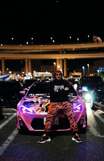 A person sitting on a pink sports car with racing decals, parked on a city street at night. 2