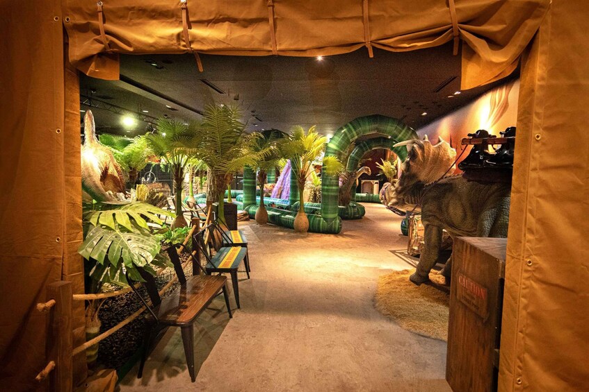 An indoor exhibit with dinosaur models, benches, and tropical plants. 2
