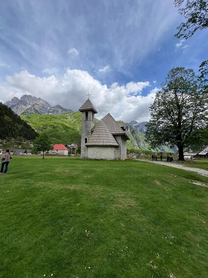 A stone church with a bell tower, surrounded by lush green grass and a large tree. 1