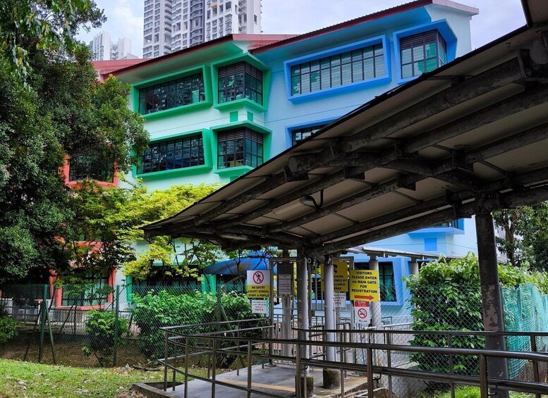A multi-story building with green and blue exteriors, surrounded by greenery and a metal fence. 2