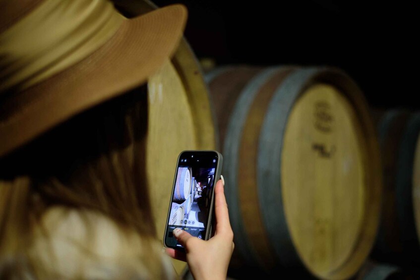 A person holding a smartphone, possibly taking a photo or video, with wooden barrels in the background. 5