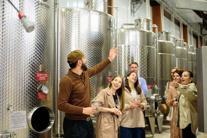 A group of people in a winery, some holding wine glasses, standing near stainless steel fermentation tanks. 1