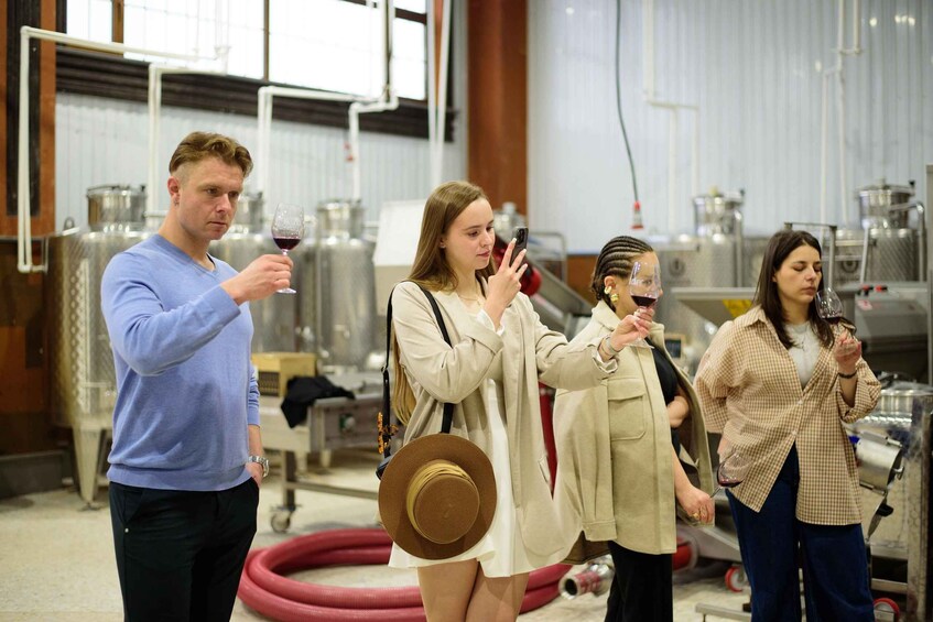 People tasting wine in a winery with stainless steel tanks and hoses in the background. 3
