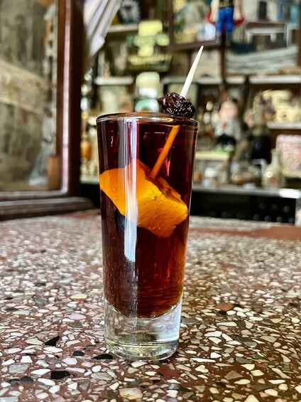 A tall glass with a dark liquid, an orange slice, and a straw on a mosaic-tiled surface. 4