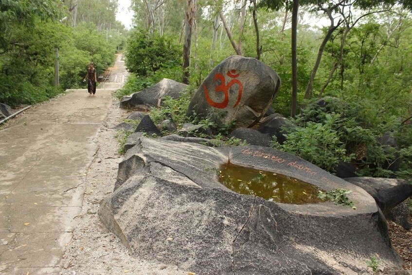 A forested path with a large rock featuring an 'Om' symbol, a small pond, and a person walking away. 2