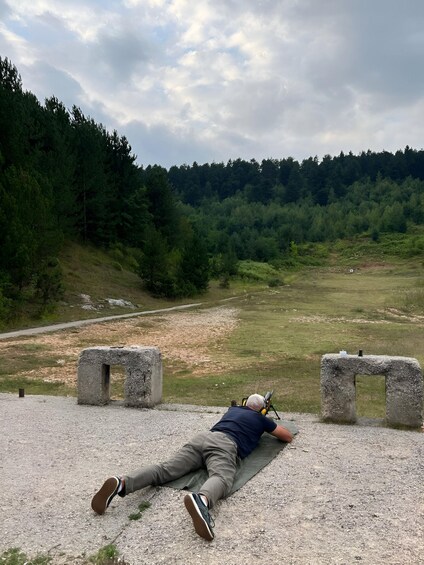 A person lying on a mat with a rifle, aiming at a distant target in a forested area. 3