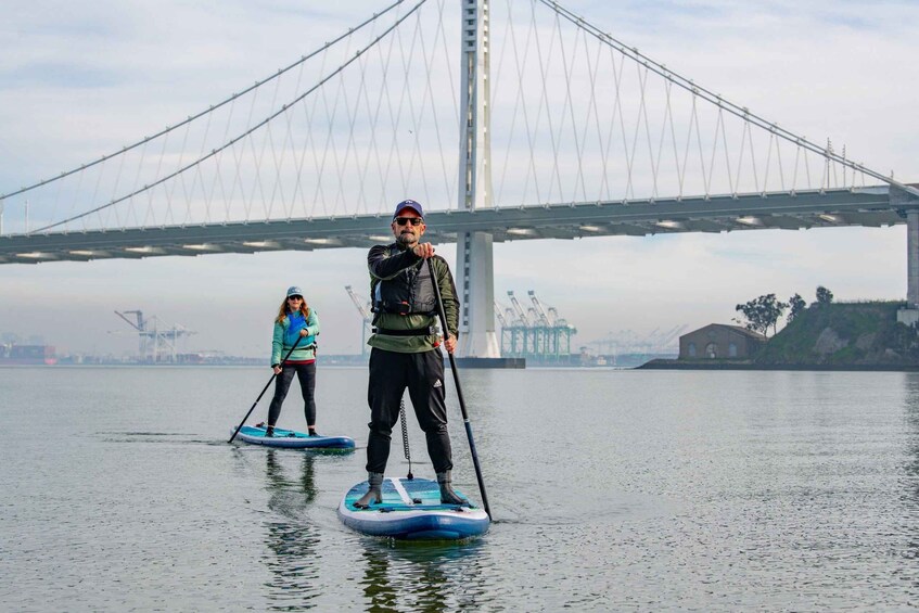 Two people paddleboarding on a calm river with a large bridge in the background. 1