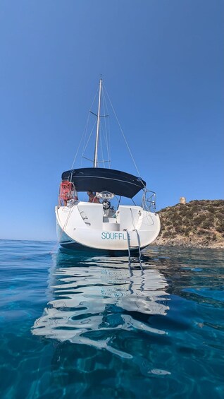 A white sailboat named 'SOUFFLE' anchored in clear blue water with a rocky coastline in the background. 1