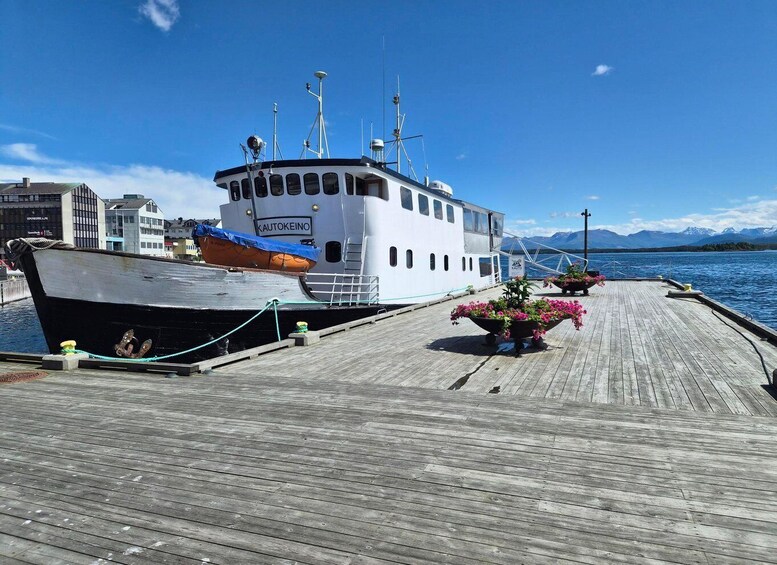 A white boat named 'KAUTOKEINO' docked at a wooden pier with flowers and a mountainous landscape in the background. 2