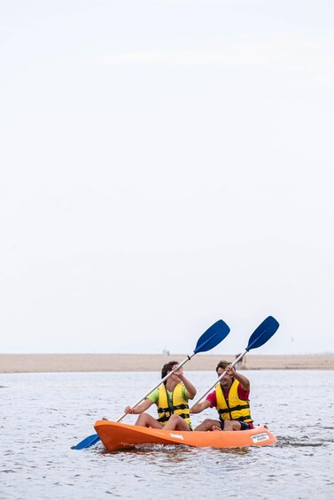 Two people in a small orange kayak with blue paddles, wearing life jackets, on a calm body of water. 1