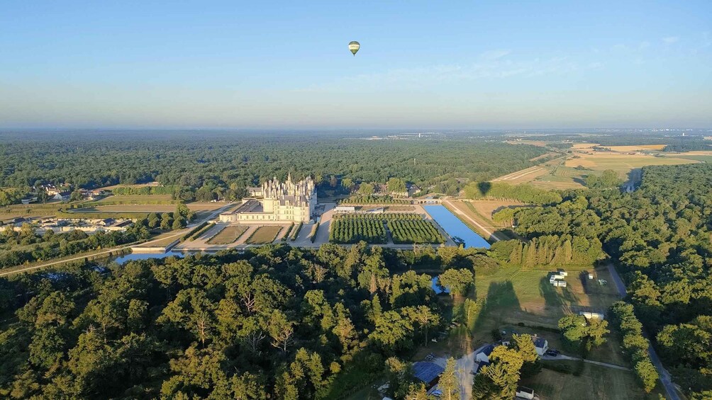 An aerial view of a castle surrounded by forested areas, a large water body, and a hot air balloon in the sky. 2