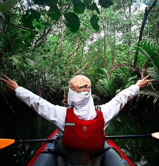 A person in a red life jacket and white shirt paddling a canoe through a dense forest. 1