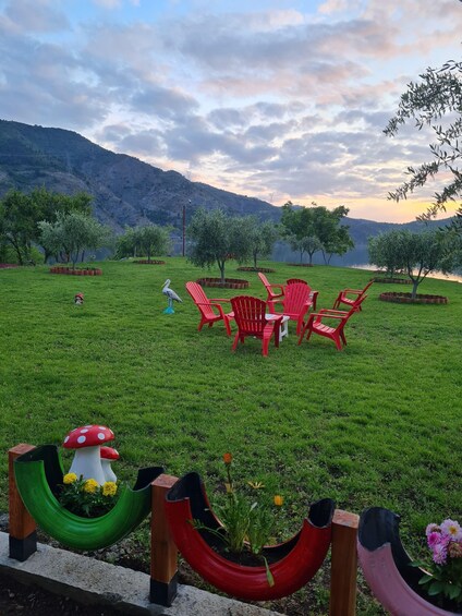A grassy area with red chairs and a table, surrounded by trees and a lake. 3