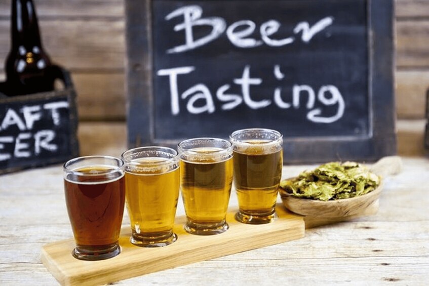 Four glasses of beer on a wooden board with a sign reading 'Beer Tasting' in the background. 2