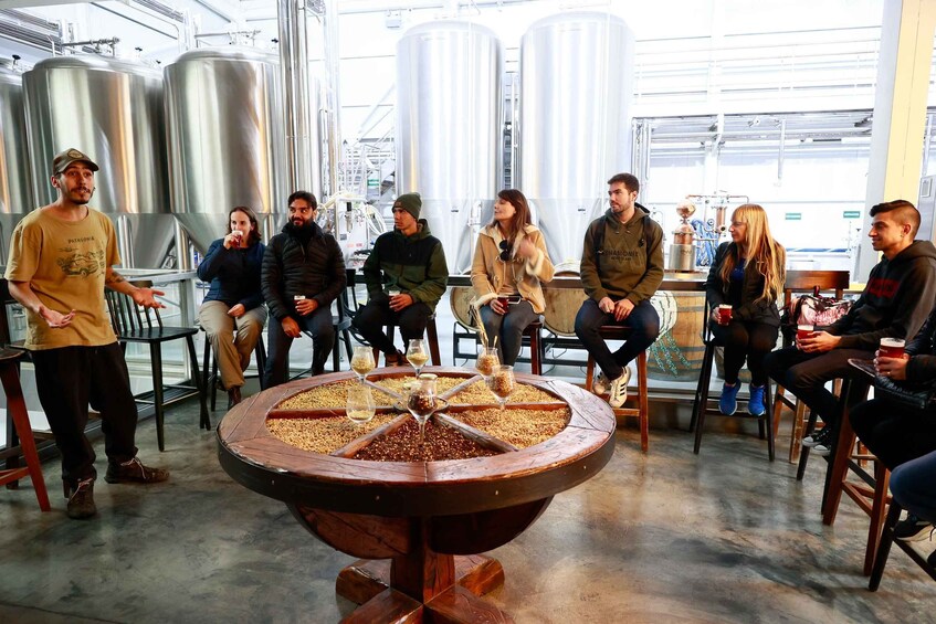 A group of people seated around a wooden table with a central grain-filled bowl, in a brewery setting with stainless steel tanks in the background. 4