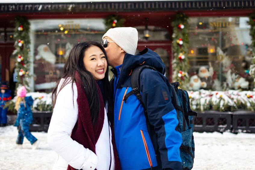 A couple embracing in a snowy setting with festive decorations and a Christmas tree visible in the background. 2