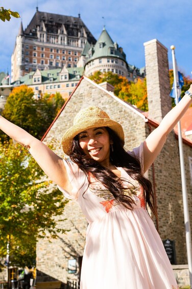 A person in a straw hat and light dress with arms raised, standing in front of a historic building with a tower and green roof. 1