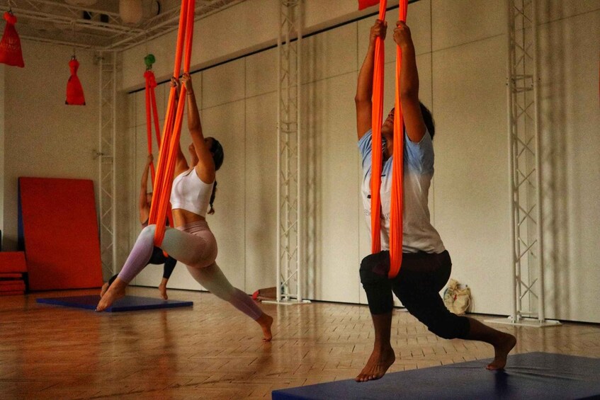 Two people performing aerial yoga with orange silks in a studio with wooden floors and red walls. 3