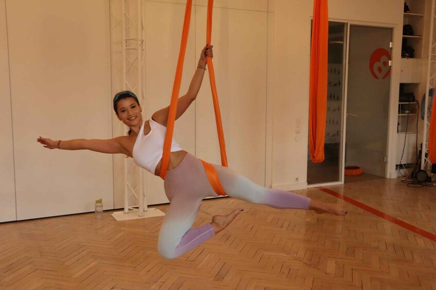 A person performing aerial yoga with orange aerial silks in a room with wooden flooring and white walls. 4