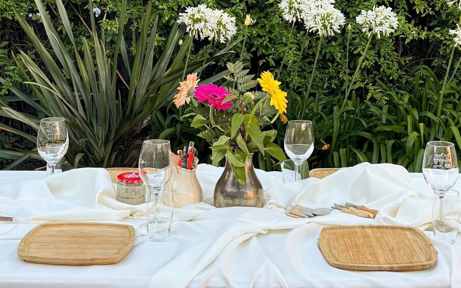 A table set outdoors with a white tablecloth, wine glasses, wooden serving boards, and a vase with flowers. 1