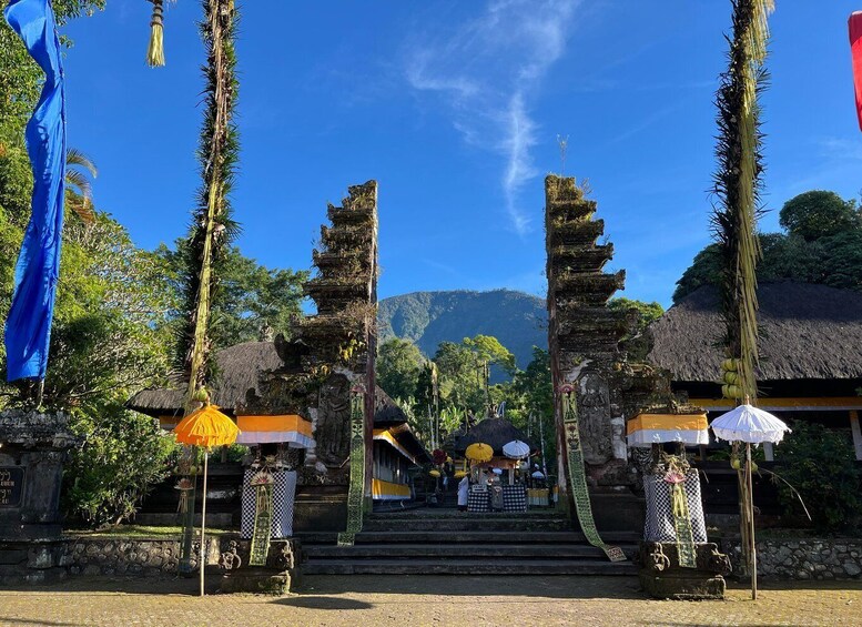 A traditional Balinese temple entrance with steps leading up to a thatched roof structure, surrounded by lush greenery and palm trees. 1