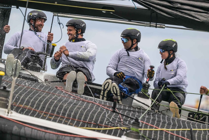 Un gruppo di persone su una barca a vela, con caschi e giubbotti di salvataggio, con corde e vele ben visibili. 3