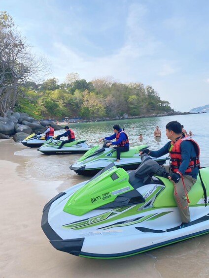 Jet skis on a sandy beach with people wearing life vests, surrounded by trees and a calm body of water. 1