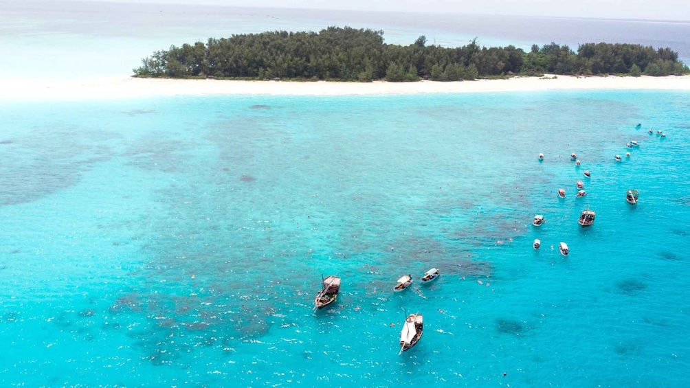 A turquoise sea with small boats anchored near a sandy beach and a forested island in the distance. 4