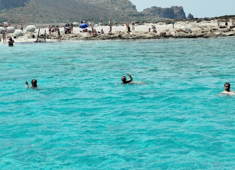 People swimming in clear turquoise water at a beach with rocky terrain and a crowd of people in the background. 3