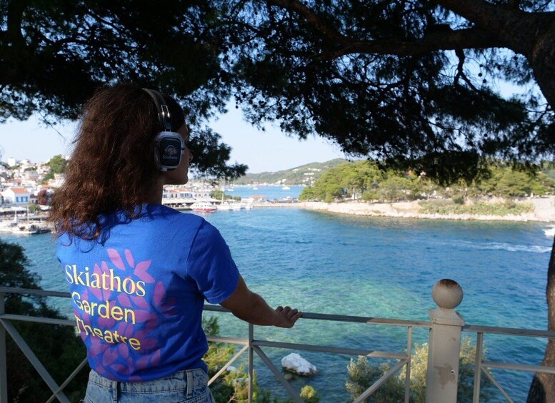 A person wearing a blue shirt with 'Skithos Garden Theatre' printed on the back, standing by a railing overlooking a coastal area with clear blue water and a town in the background. 3