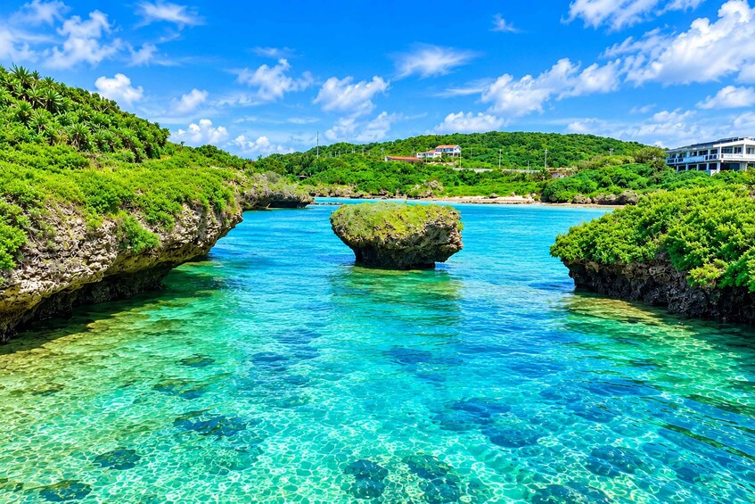 A clear turquoise river with a central rock formation surrounded by lush greenery and a distant building. 3