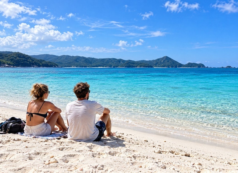 Two people sitting on a sandy beach facing a turquoise sea with clear skies and distant green hills. 2