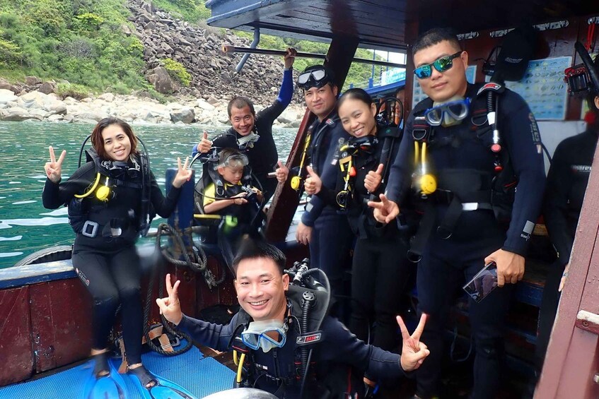 A group of people in scuba diving gear on a boat, with rocky terrain and water in the background. 1