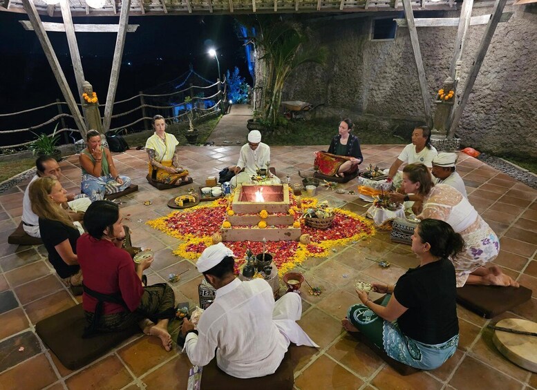 A group of people seated in a circle around a central fire pit, with a stone wall and a rope barrier in the background. 2
