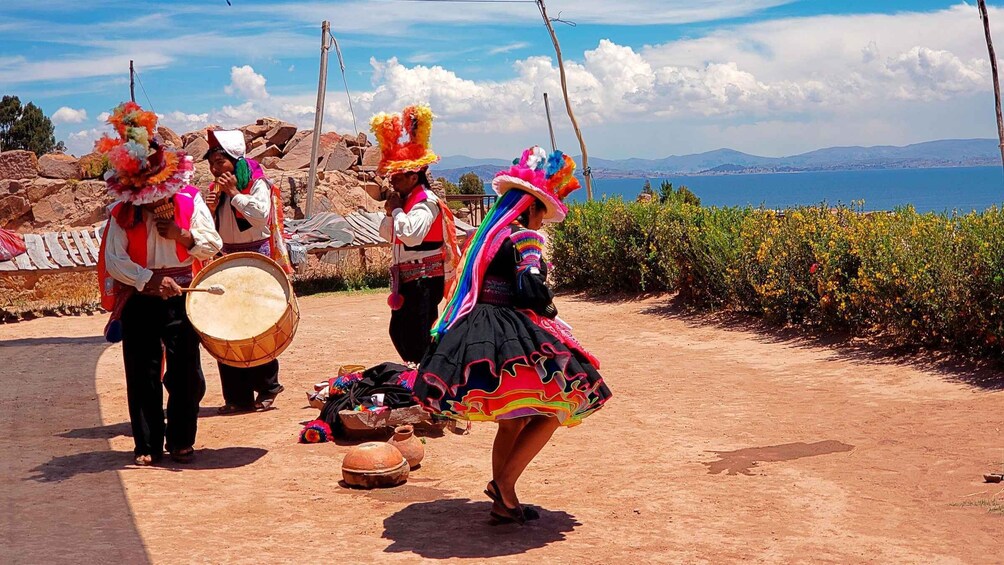 A group of people in traditional attire performing a dance with drums, set against a coastal landscape with clear skies. 5