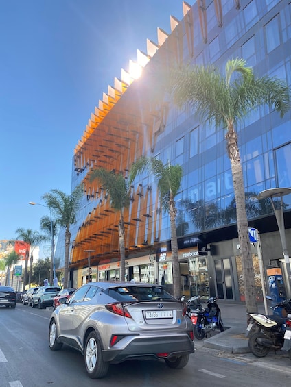 A modern building with reflective glass windows, palm trees, and a silver car parked on the street. 3